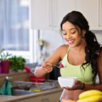 Smiling woman eating a nutritious fruit bowl in a bright kitchen setting.